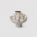 A sculptural ceramic vase with a textured gray base and a top composed of irregular, petal-like off-white glazed shards outlined in dark glaze, photographed on a light gray background.