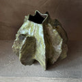 A sculptural, organic-shaped ceramic vase with a textured, leaf-like surface in earthy olive green, gray, and brown tones, set against a neutral, warm-toned background.