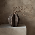 A dark textured ceramic vase with a sculptural, ribbed form holding a gnarled decorative branch, displayed on a concrete pedestal against a textured plaster wall.