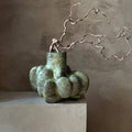 An organic-shaped green ceramic vase with a textured matte finish, holding a cluster of wavy branches, displayed on a light beige pedestal against a textured brown plaster wall.