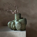 An abstract, organic-shaped ceramic vase in textured sage green sits on a light concrete pedestal against a warm, textured plaster wall, styled with a single thin, branching twig.