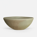 A handmade ceramic bowl with a wide, shallow profile, featuring a textured matte glaze in a muted sage green with earthy brown undertones, set against a clean white background.