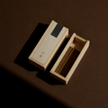 A light natural wood incense box shown open, with incense sticks inside, on a dark brown surface under dramatic directional lighting.