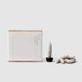 White rectangular card with wooden border and text, next to a small black pot with a candle and additional candles on a white background.