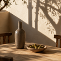 A rustic matte gray ceramic bottle stands on a wooden table next to a shallow wooden bowl filled with green olives, set against a sunlit stucco wall with tree branch shadows.