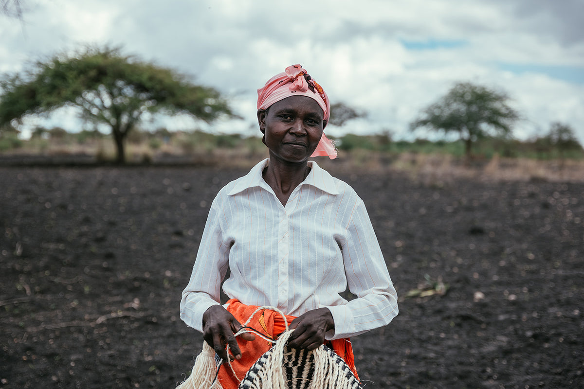 A weaver from Kitui holds a basket they made. Black dirt and trees are sat behind them.