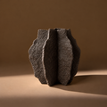A dark textured ceramic vase with a sculptural, ribbed organic shape, photographed on a warm brown background with dramatic side lighting.