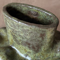 Close-up of a ceramic vessel with a rounded, bulbous form and a glossy glaze in olive green, brown, and beige tones. A small embossed monogram logo is visible inside the opening against a warm beige background.