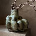 An abstract sculptural vase with a bulbous organic shape in mottled olive green and gray, holding a twisted brown branch-like stem, set on a stone pedestal against a textured warm brown wall.