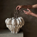 A sculptural ceramic planter with a textured, layered organic shape in warm off-white, displayed on a concrete pedestal against a dark brown plaster wall. 