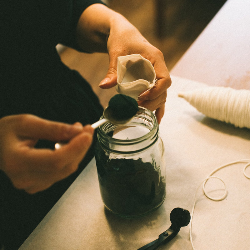 Person using a spoon to scoop something from a jar on a table with various items.