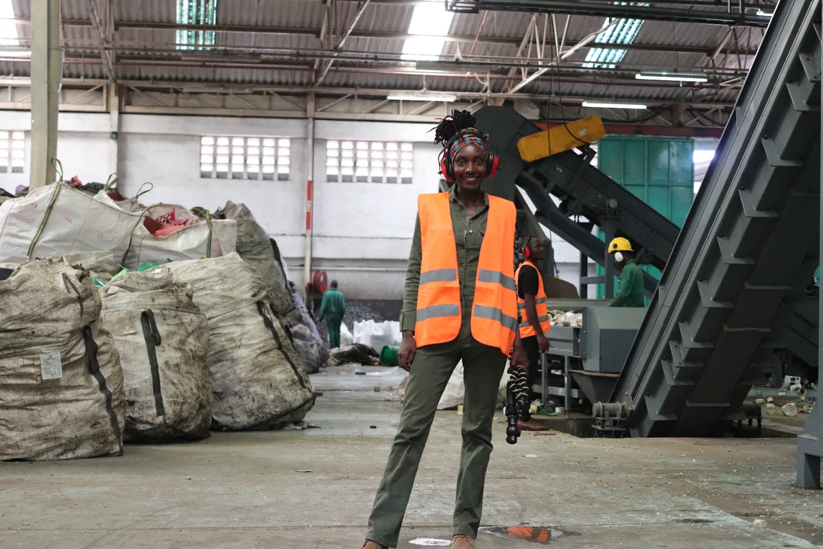 A person standing in a large industrial recycling facility, wearing a bright orange safety vest over dark clothing, with large white bags, machinery, and other workers visible in the background.