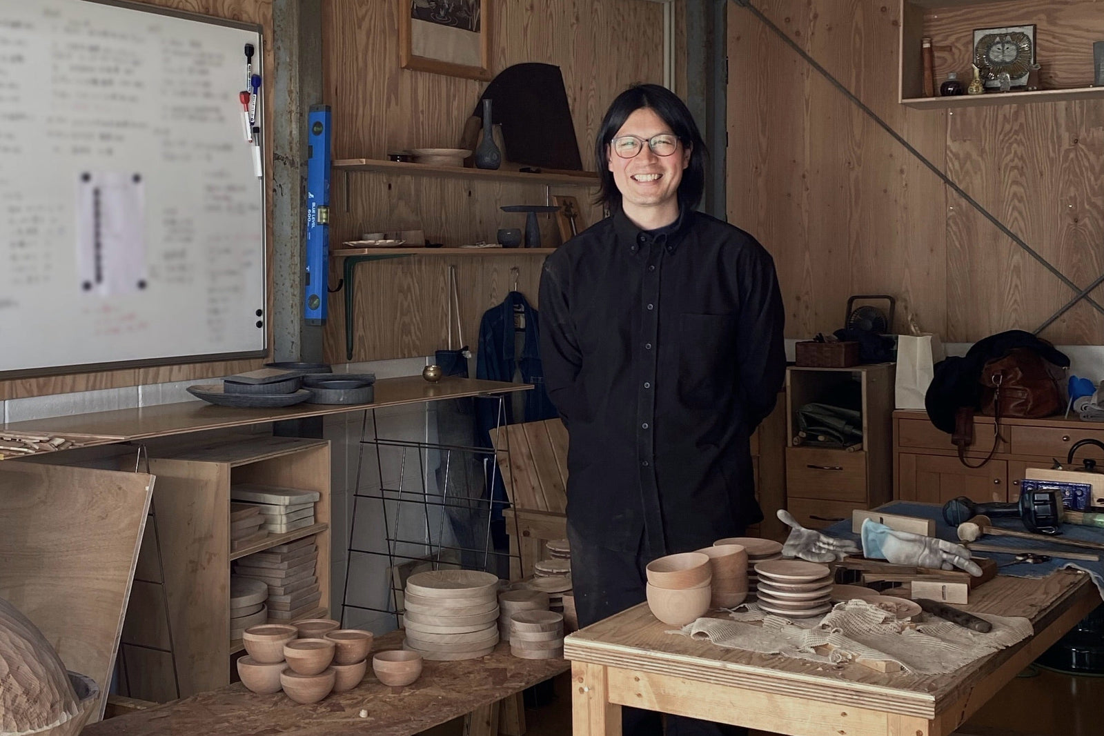 Kinji Yoshida, Obakki artisan partner, wearing a black long-sleeve top stands in a wood-paneled pottery studio with tables, tools, and ceramic bowls.