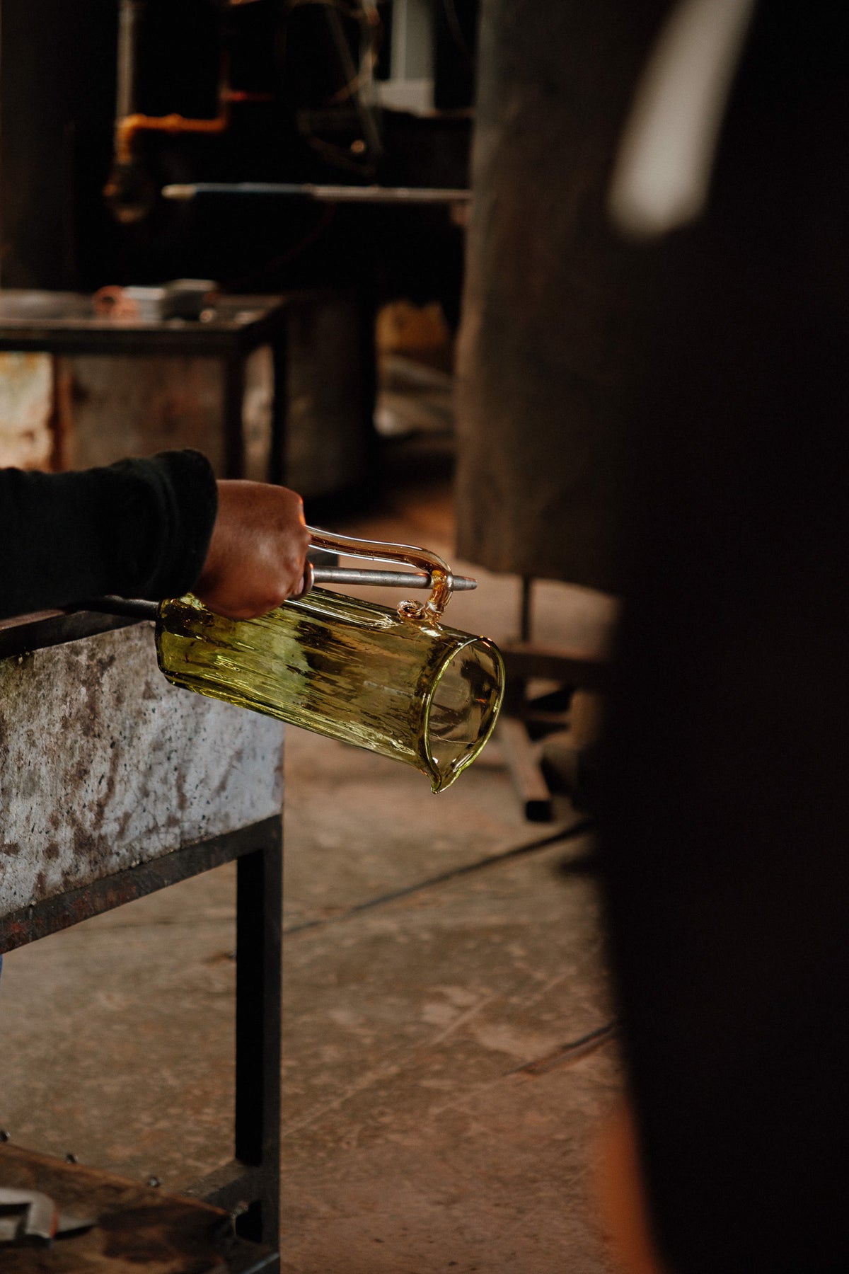A hand holding a translucent olive-green glass pitcher in a workshop setting with a blurred background.