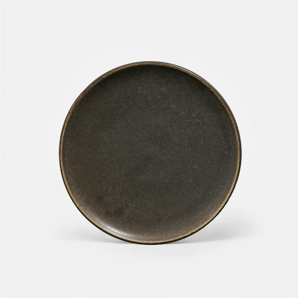 A round ceramic plate with a dark, matte charcoal-brown glaze and a contrasting warm brown rim, photographed from a top-down angle on a clean white background.