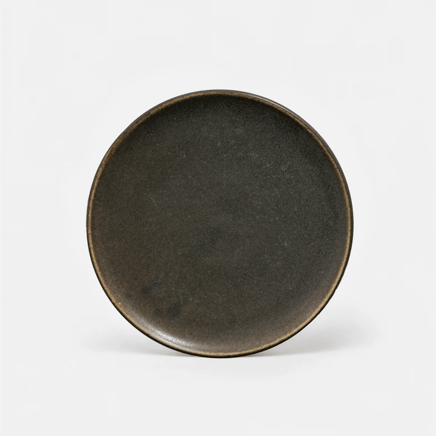 A round ceramic plate with a dark, matte charcoal-brown glaze and a contrasting warm brown rim, photographed from a top-down angle on a clean white background.