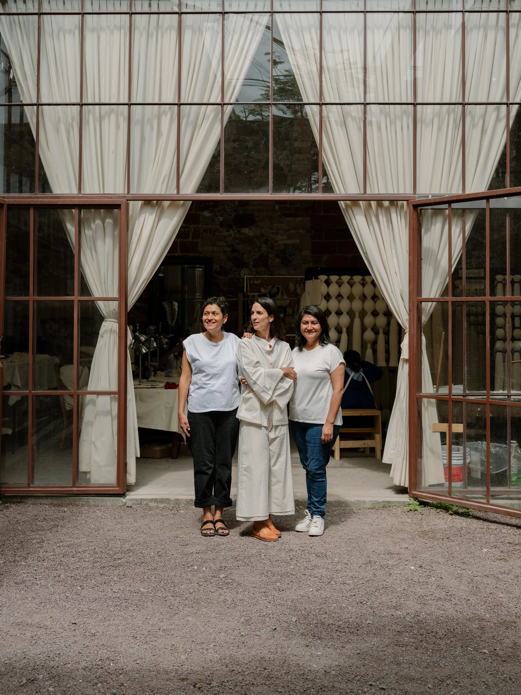 Three women standing in front of large glass studio doors at the Caralarga workshop in Mexico, artisans working with Obakki.
