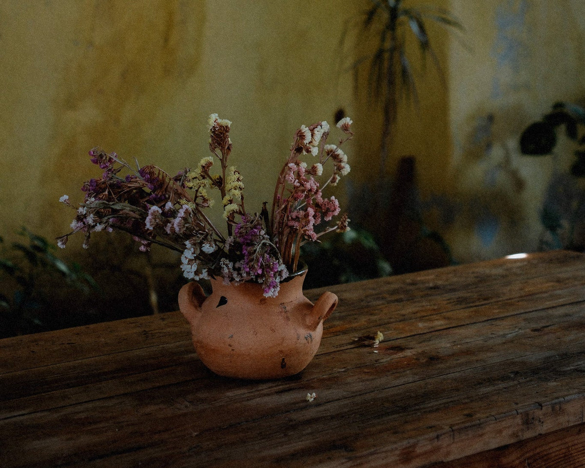 A small terracotta clay flower pot filled with dried pink and white flowers, placed on a rustic wooden table against a textured yellow wall.
