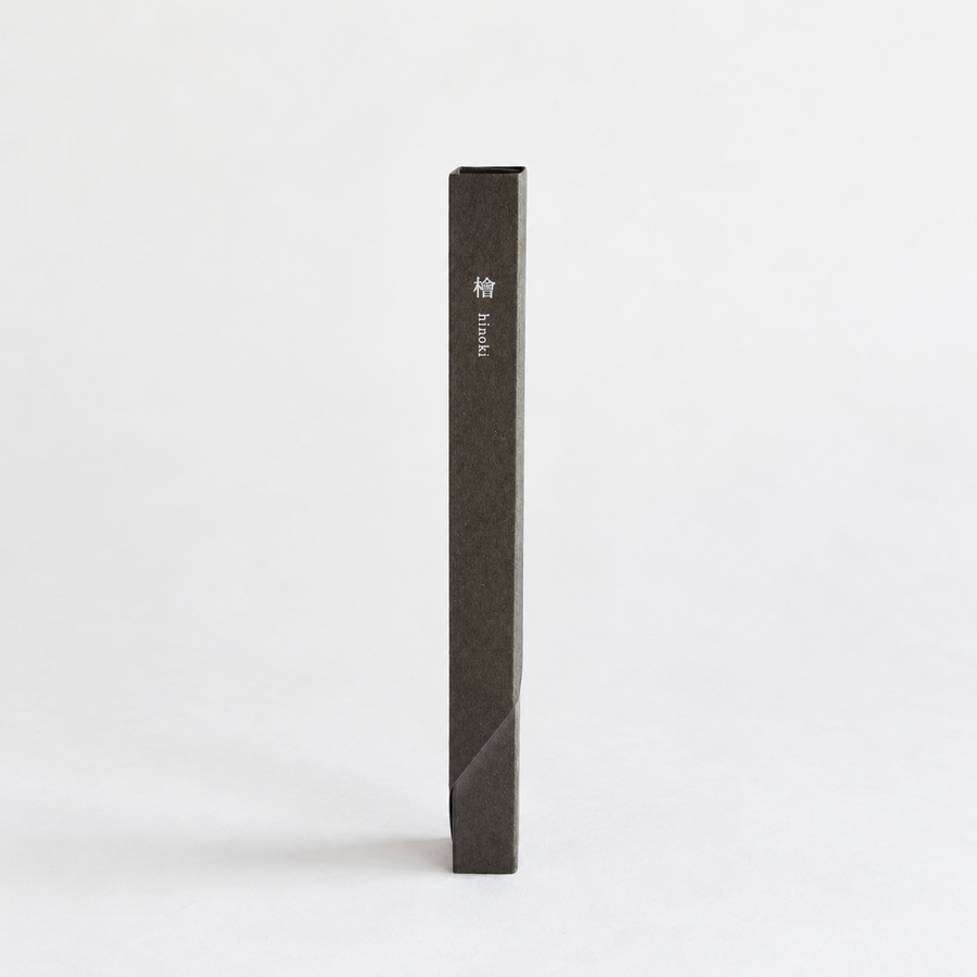 A tall, narrow dark charcoal gray incense box standing vertically on a light gray background. The box has small white text printed on the side in both English ('Hinoki') and Japanese characters.