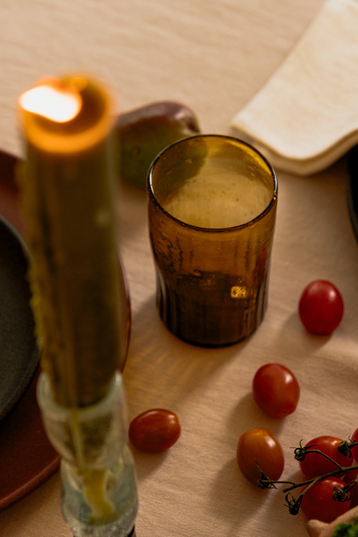 An amber glass candle holder or tumbler with a lit candle inside, styled on a beige tablecloth with cherry tomatoes, a dark plate, and a folded cream napkin.