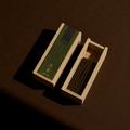 A wooden incense box shown open with incense sticks inside, alongside the matching lid. The lid features a dark green label with concentric circle patterns and gold Chinese characters reading 'Agarwood' and '沈香'.