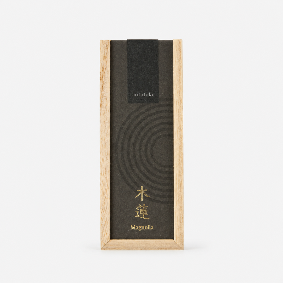 A tall rectangular incense box with a light natural wood frame and a dark charcoal lid featuring concentric circle lines and gold text reading 'hitotoki' and 'Magnolia'.