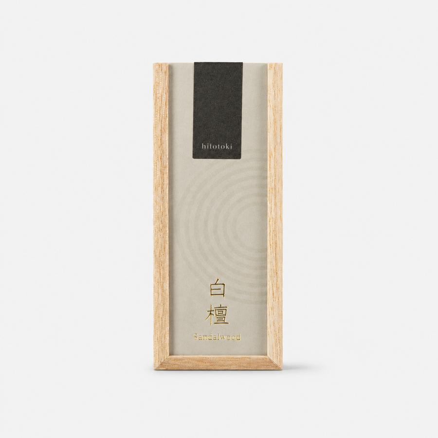 A tall rectangular incense box with a light natural wood frame and a cream-colored lid featuring a subtle circular pattern, a black label near the top reading 'hitotoki', and gold text near the bottom reading 'Sandalwood', shown on a clean white background.