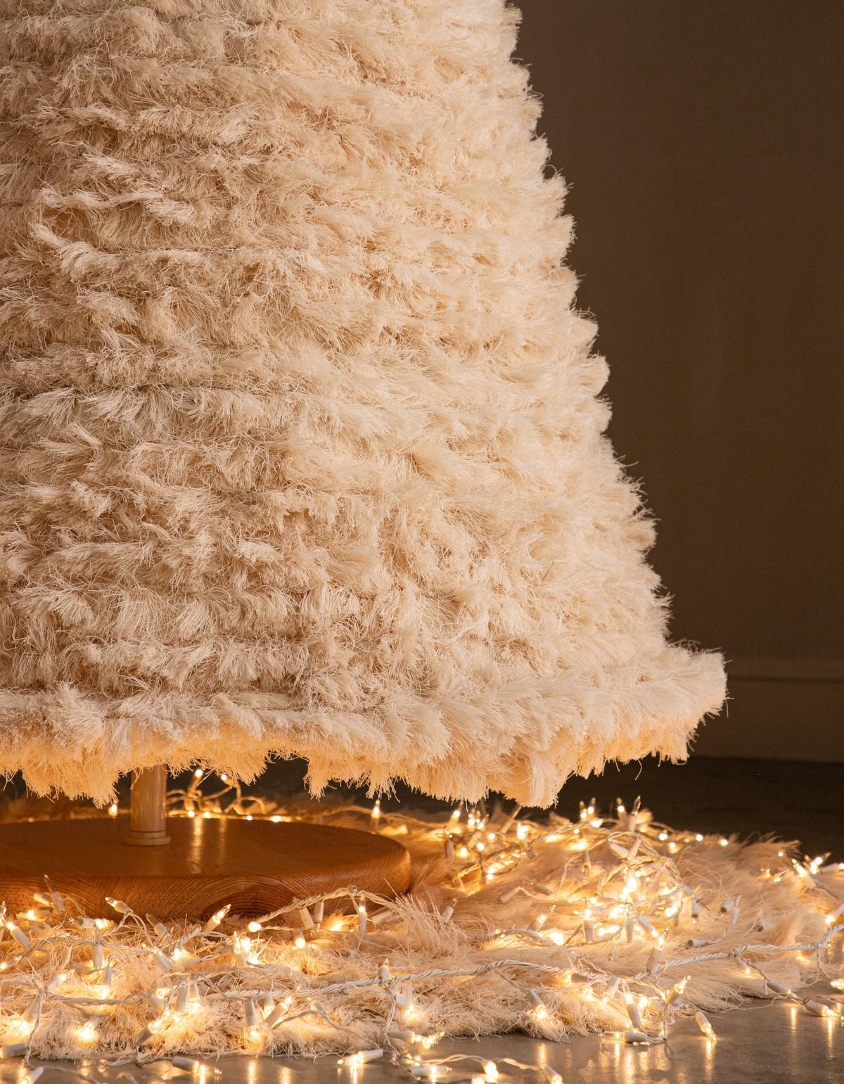 A close-up of a fluffy, shaggy cream-colored Christmas tree with warm white string lights glowing around its base against a dark brown background.