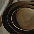 A stack of ceramic plates in earthy tones, including a large dark brown plate, a medium brown plate, and a light beige plate, arranged on a neutral fabric surface with dramatic side lighting.