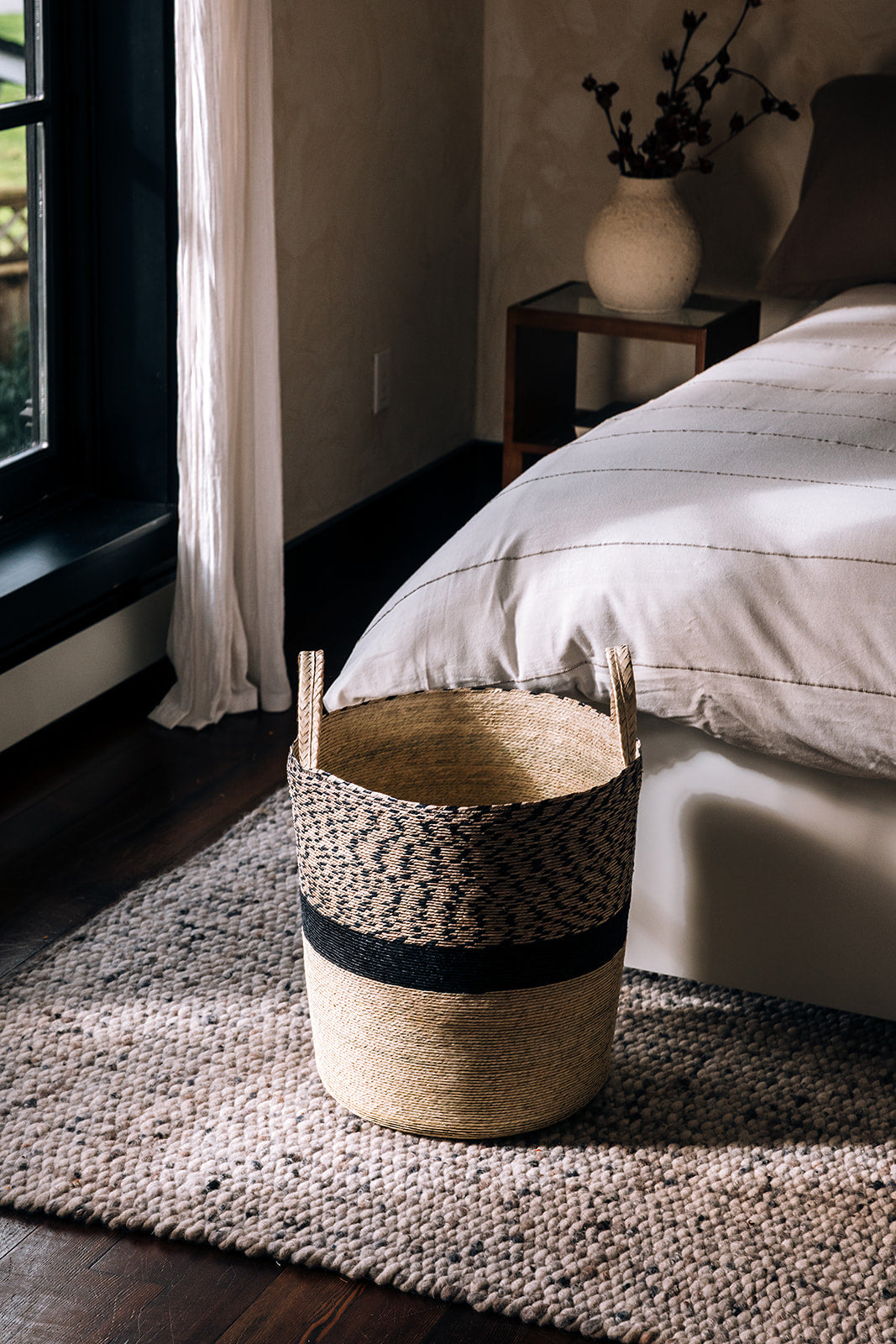 Basket sits in front of a bed. Sunlight shines through the window.