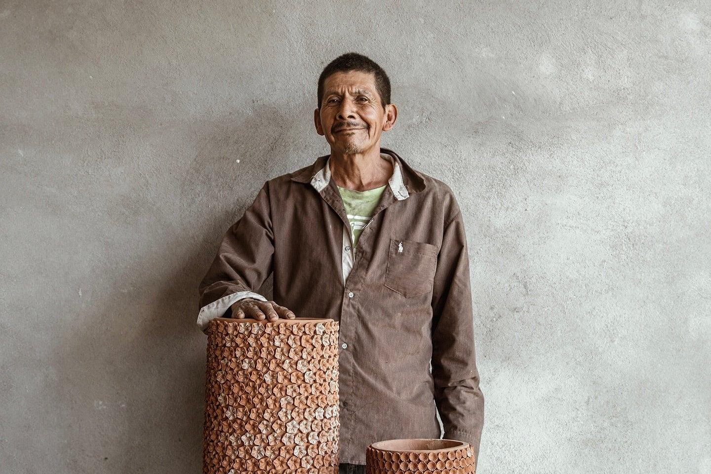 A man wearing a brown work shirt stands behind three tall, textured terracotta planters with a circular shell-like pattern, set on a rustic wooden table against a gray concrete wall.