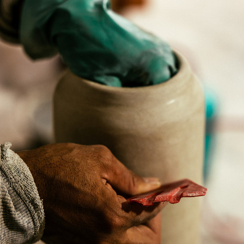 Person wearing green gloves and holding a cylindrical clay object with a blurred background