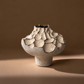 A sculptural ceramic vase with a textured, organic cracked surface and a short cylindrical base, shown on a warm brown studio background with a soft shadow.
