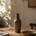 A rustic brown ceramic vase filled with dried baby's breath stems, set on a wooden table in a warmly lit room with a bowl and framed wall art in the background.