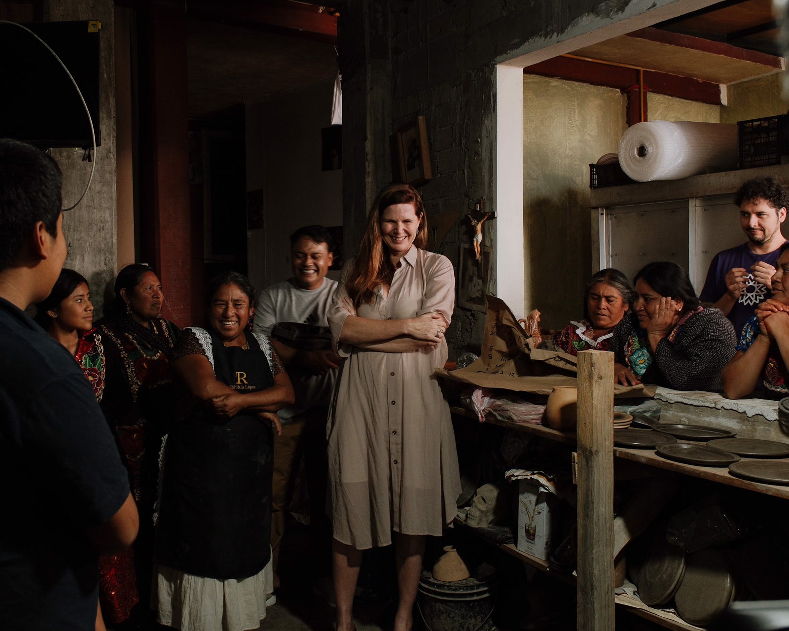Treana and Obakki's artisan partner Rufina in a rustic indoor workshop or kitchen, surrounded by several women and shelves of pottery on a wooden table.