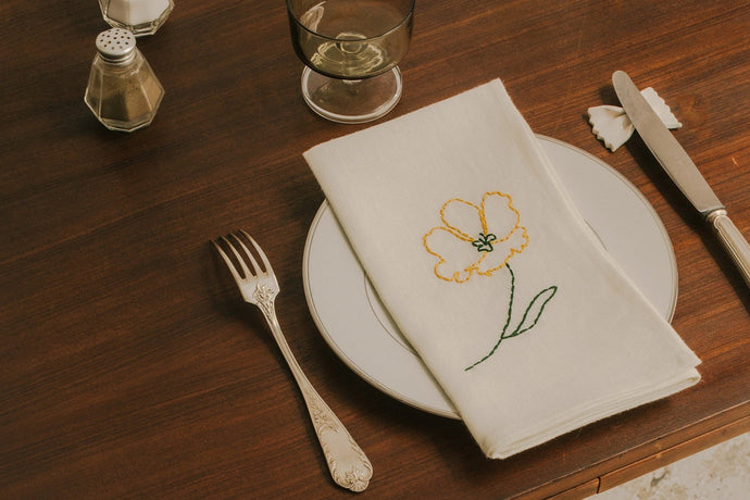 Dining table setting with embroidered napkins, glasses, and cutlery on a wooden surface.