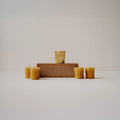 A set of six natural beeswax taper candles in a warm golden-yellow hue, arranged symmetrically around a central rectangular wooden plaque with engraved text, on a seamless light beige studio background.