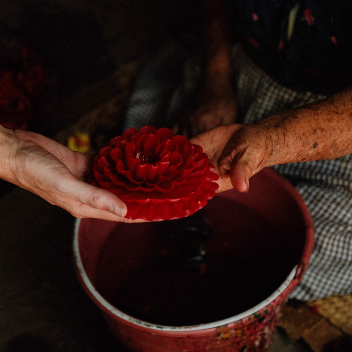Viviana holds a carved red paper lantern shaped like a flower above a red bucket, with more red lanterns visible in a woven basket at the bottom. The scene is photographed in a dim, rustic setting with shallow depth of field.
