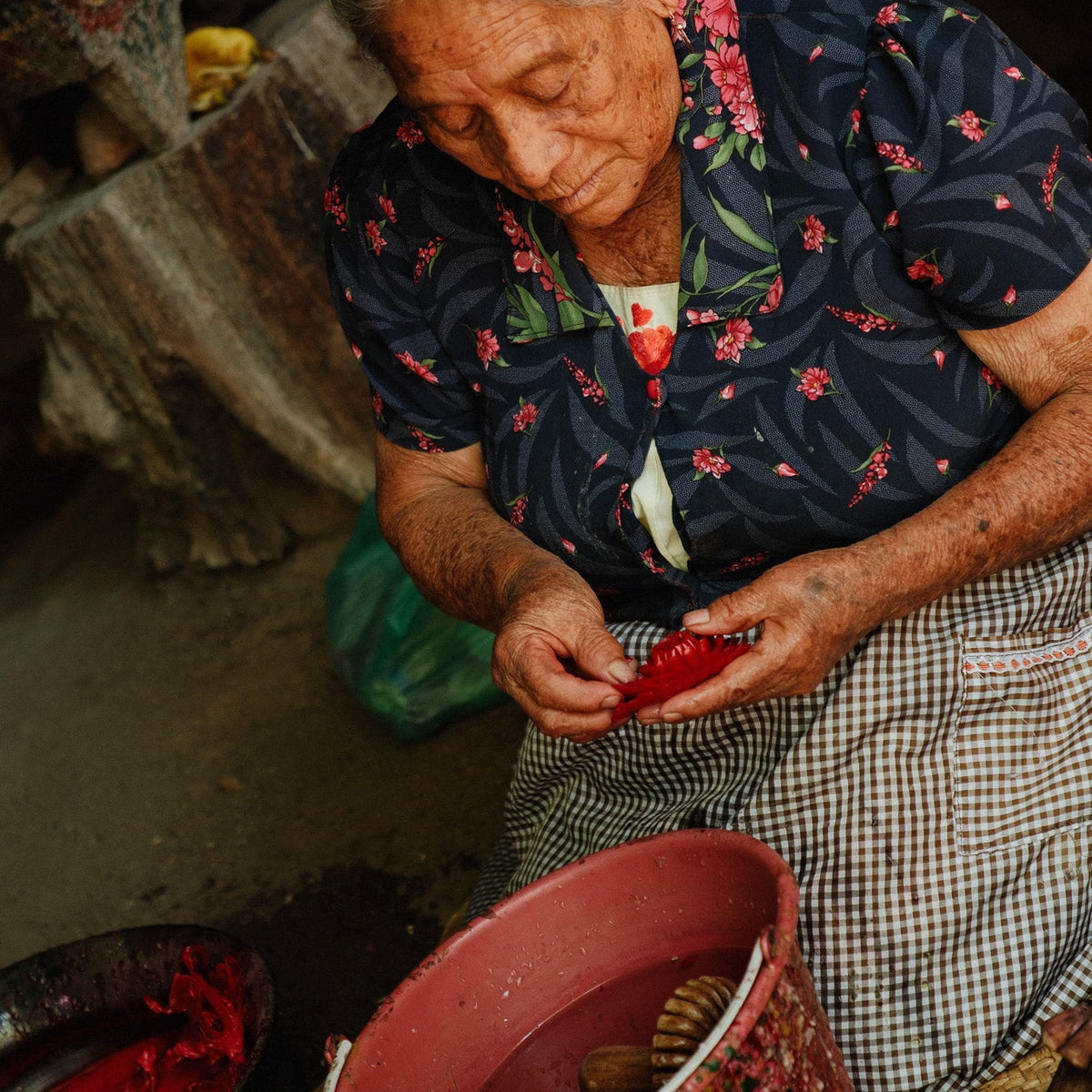 Viviana in a floral navy blouse and checkered skirt sits on the floor, handling red material in a rustic setting with buckets and pottery nearby.