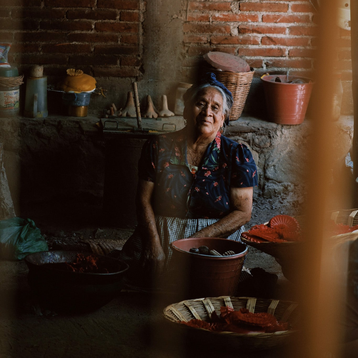 An elderly woman with a blue headwrap sits in a rustic room with exposed brick walls, surrounded by cooking pots, baskets, and wooden tools, viewed through a blurred foreground frame.