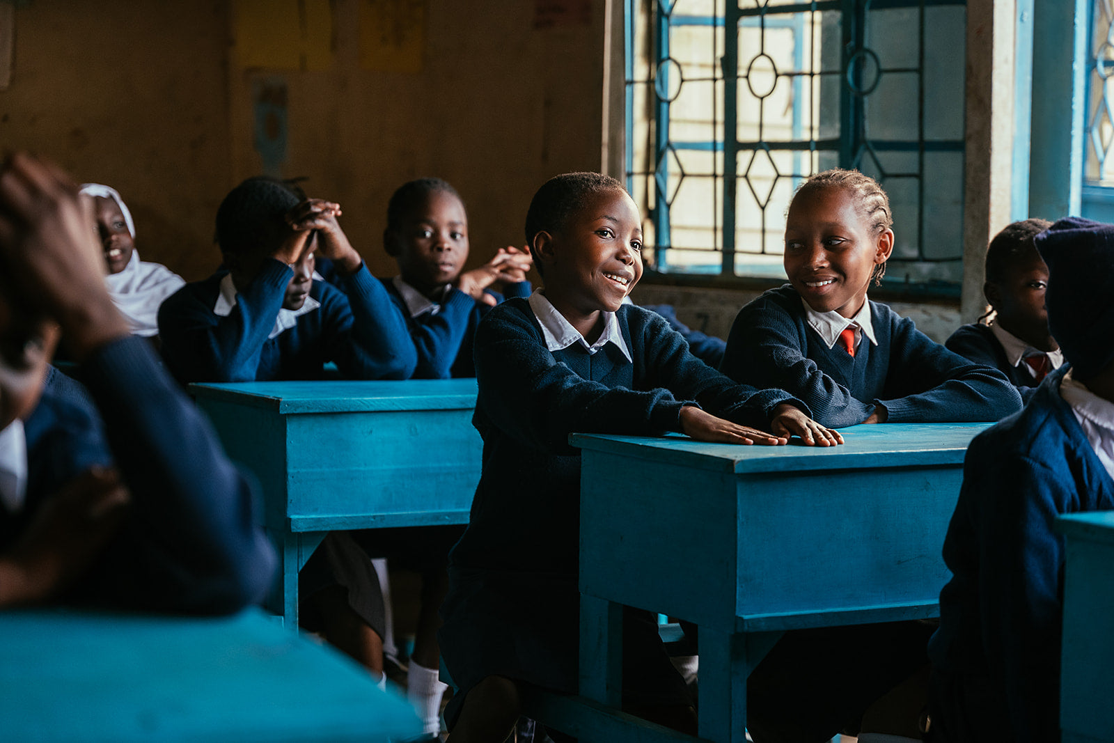 Children sitting at blue desks in a classroom with a window in the background.